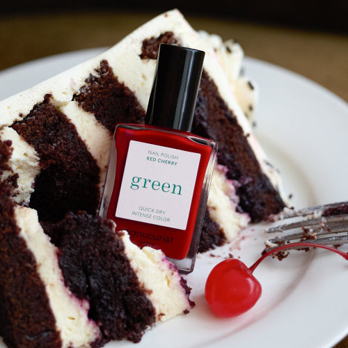 A bottle of red nail polish labeled Red Cherry sits in front of a slice of chocolate cake with white frosting and a maraschino cherry, with a fork resting nearby.