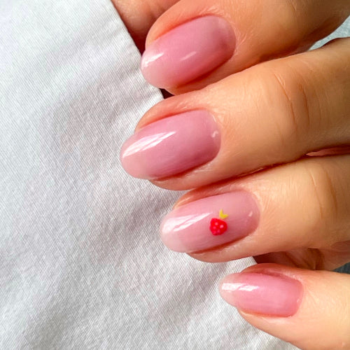 A hand with neatly manicured nails painted in a glossy, light pink shade. One nail features a small red strawberry design with a yellow stem.