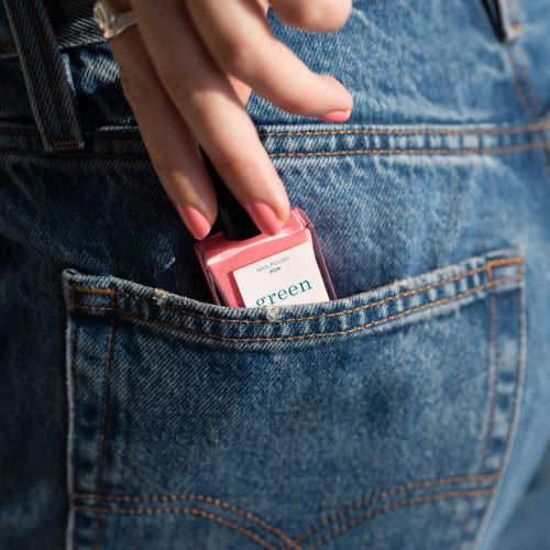A hand places a bottle of pink nail polish labeled green into the back pocket of blue denim jeans.