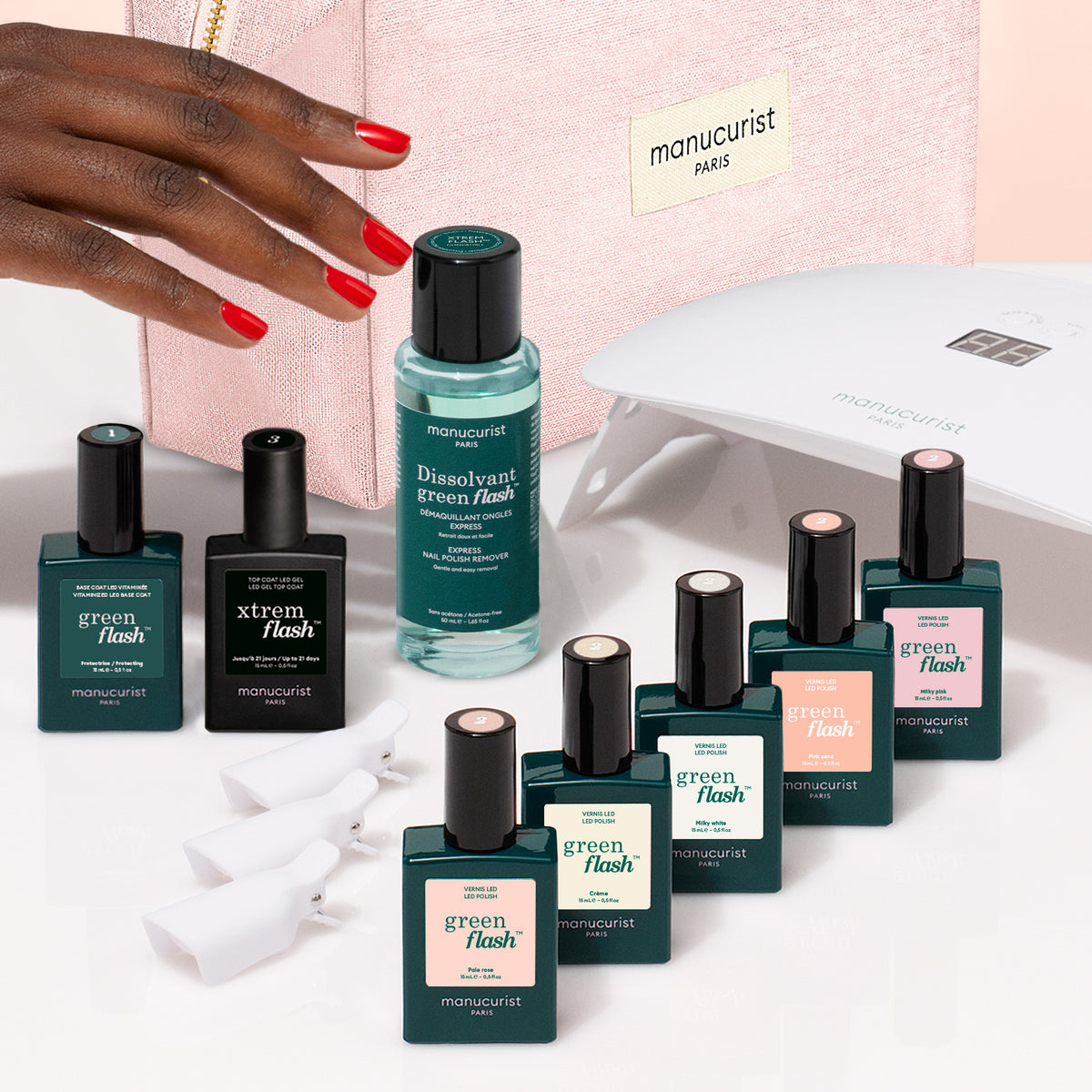 Une main aux ongles rouge vif surplombe un Super Starter Kit Manucurist de vernis semi-permanent LED Soap Nails, comprenant une lampe LED, du dissolvant et sept flacons colorés disposés devant une trousse de maquillage rose pour une manucure ultra-brillante.