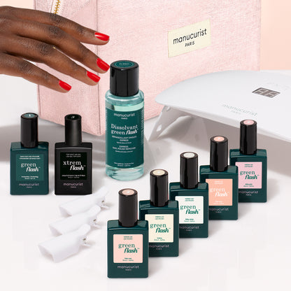 Une main aux ongles rouge vif surplombe un Super Starter Kit Manucurist de vernis semi-permanent LED Soap Nails, comprenant une lampe LED, du dissolvant et sept flacons colorés disposés devant une trousse de maquillage rose pour une manucure ultra-brillante.