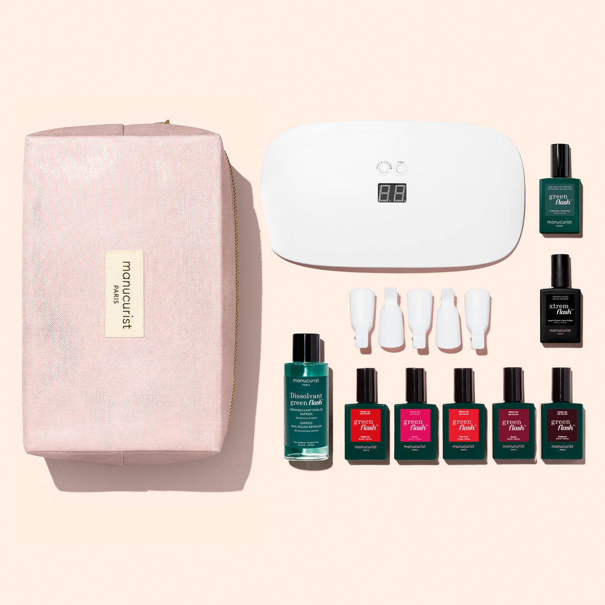 A hand with red nails rests near bottles from the Manucurist Super Starter Kit, including gel polishes, nail polish remover, a UV lamp, removal clips, and a pink bag. The products are arranged on a white surface, promising an ultra-glossy manicure that lasts.