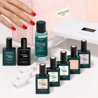 Une main aux ongles vernis de rouge se tend vers un Super Starter Kit de vernis semi-permanent LED Soap Nails, avec des flacons, du dissolvant, une lampe LED et des pinces disposés devant une trousse de maquillage rose pour réaliser une manucure ultra-brillante.
