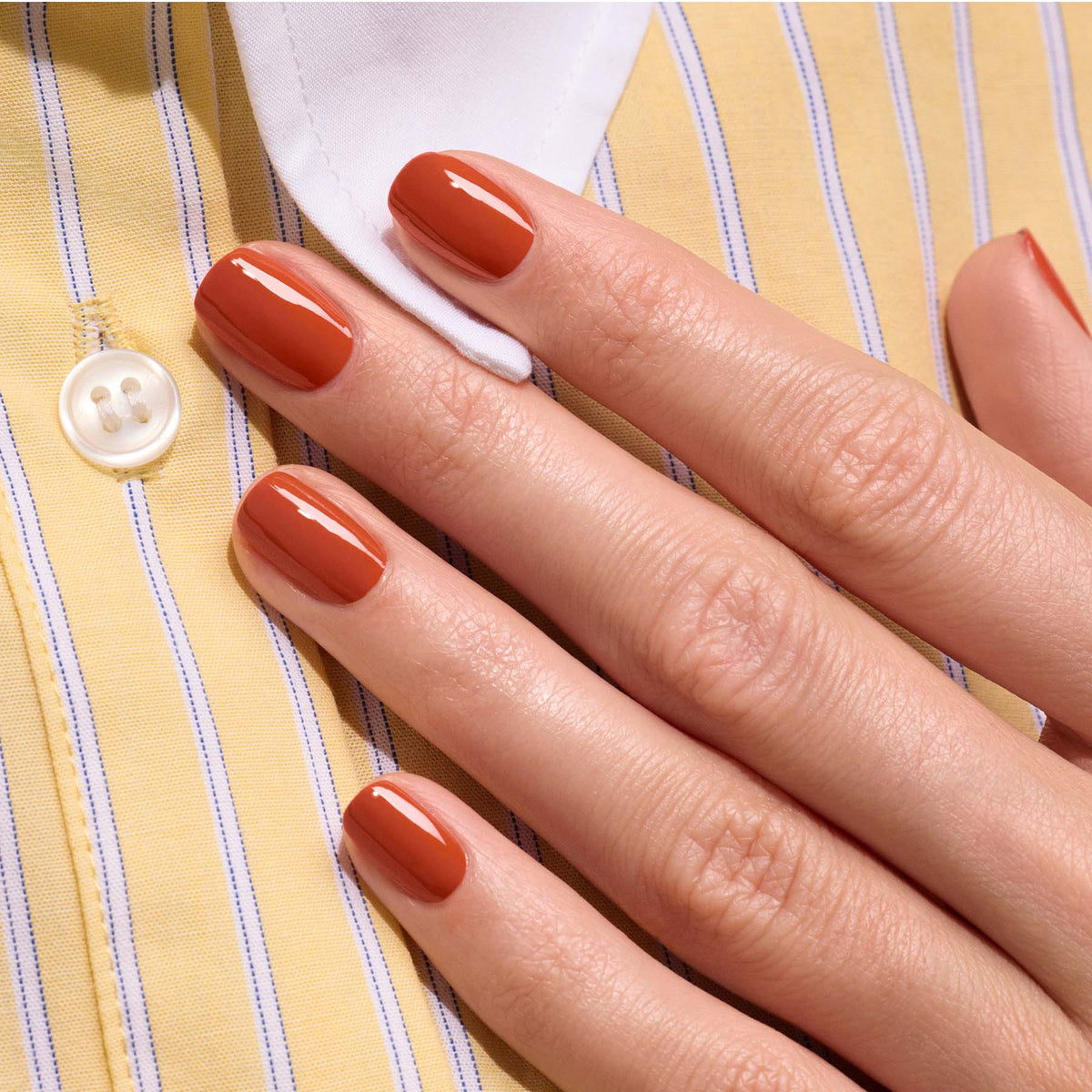 Une main aux ongles soigneusement manucurés vernis en Terracotta de Manucurist, une teinte terre brûlée brillante de la gamme de vernis gel Green Flash, repose sur une chemise à rayures jaunes et blanches avec un bouton blanc.