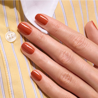 A hand with neatly manicured nails painted in Manucurist’s Terracotta, a glossy scorched red earth shade from the Green Flash gel polish range, rests on a yellow and white striped shirt with a white button.