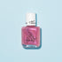 A clear bottle of Manucurist’s “Betty the Turtle” raspberry glitter pink kids’ nail polish with a white cap and turtle illustration, set on a light blue background. Easily removable with soapy water for simple cleanup.