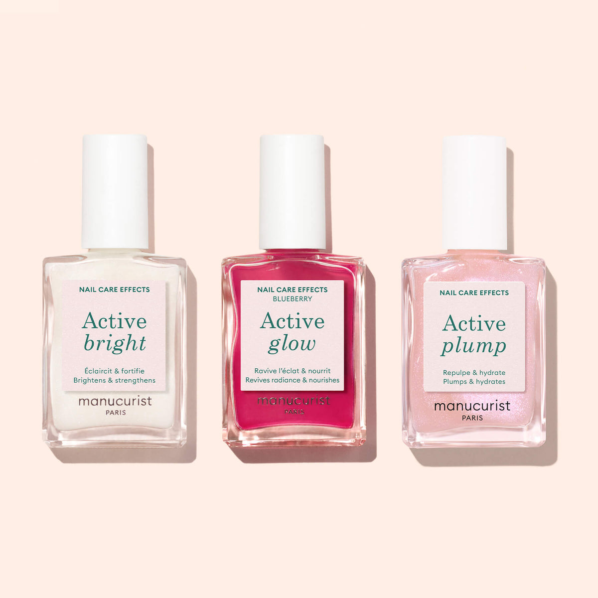 Three bottles of Manucurist nail care—Active™ Bright, Active Glow, and Active Plump—stand side by side on a light pink surface. Each bottle features a different colored liquid and white labels, highlighting this collection of bio-based nail polish and nourishing nail strengthener formulas from Manucurist.