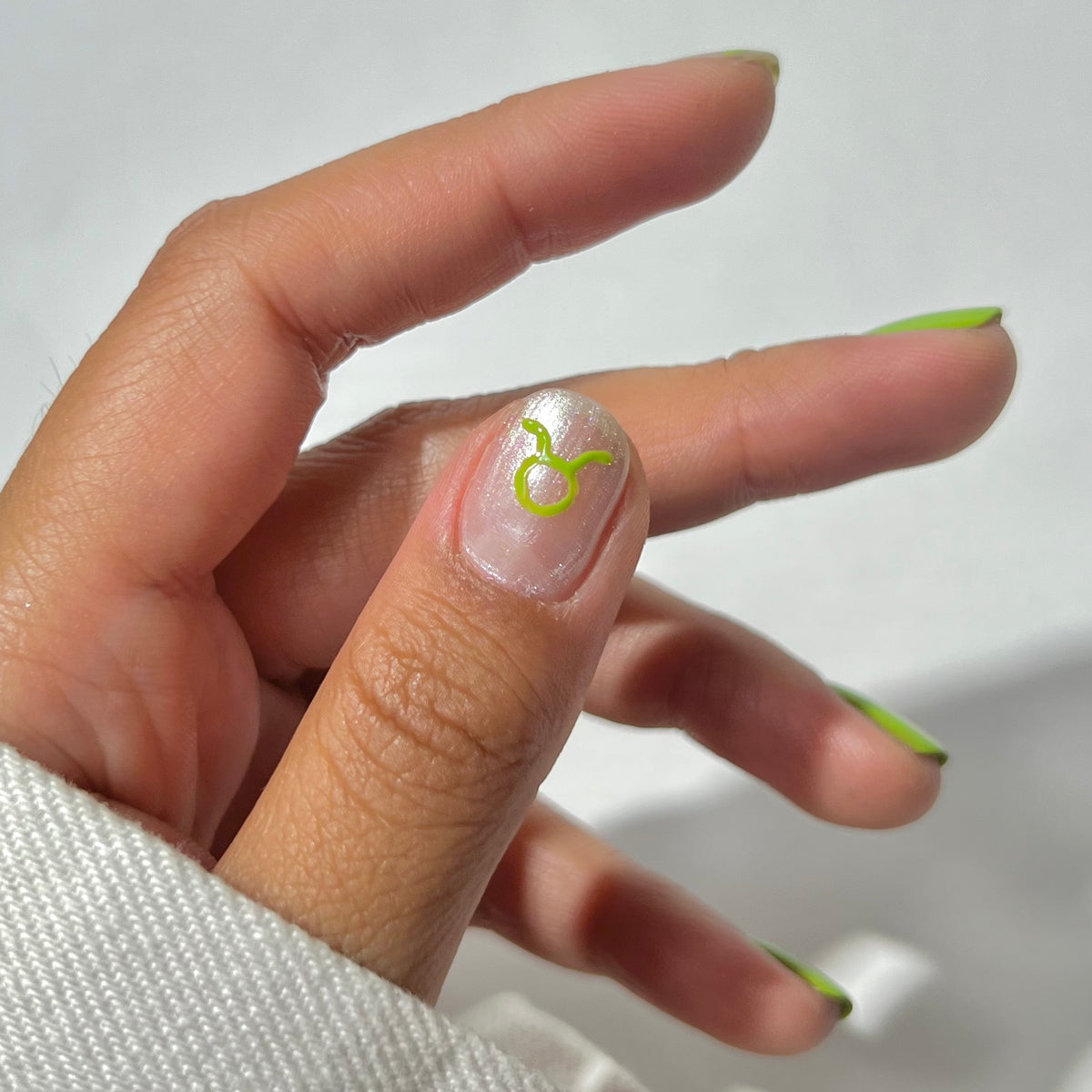 A hand with light brown skin shows nails painted with green French tips; the ring finger features a metallic silver base with a green Taurus zodiac symbol. The person wears a white long-sleeve garment.