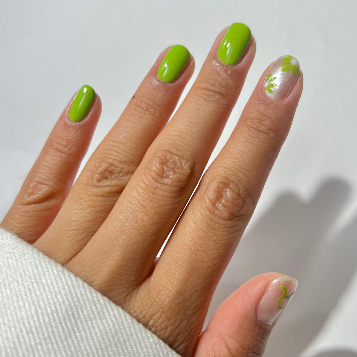 A hand with lime green nail polish on four fingers and a clear accent nail featuring green and white floral nail art. The hand is partially covered by a white sleeve.