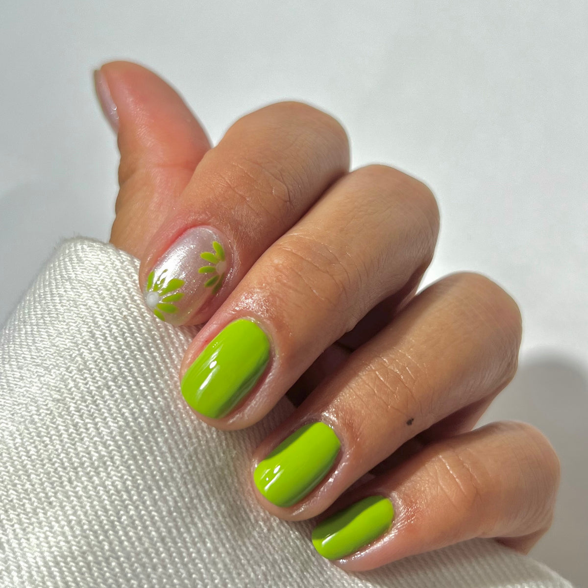 A hand with short nails painted bright green, except the thumb and index finger, which have a clear base and green leafy designs. The hand rests on a white textured fabric.