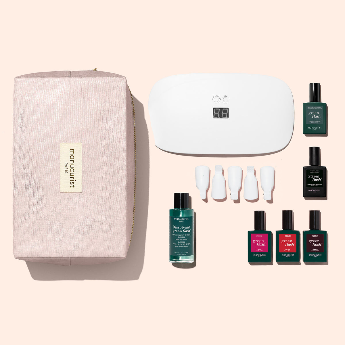 The Manucurist Starter Kit includes everything you need for an ultra-glossy manicure at home: a pink pouch, UV nail lamp, assorted nail polish bottles in various colors, vitaminized base coat, remover bottle, nail file, and four white sample sticks—all arranged neatly on a light pink background.