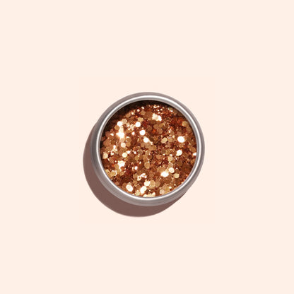 Une boîte métallique ronde de paillettes biodégradables Copper de Manucurist révèle des reflets dorés et bronzés sur un fond beige clair, projetant une douce ombre sur la gauche.