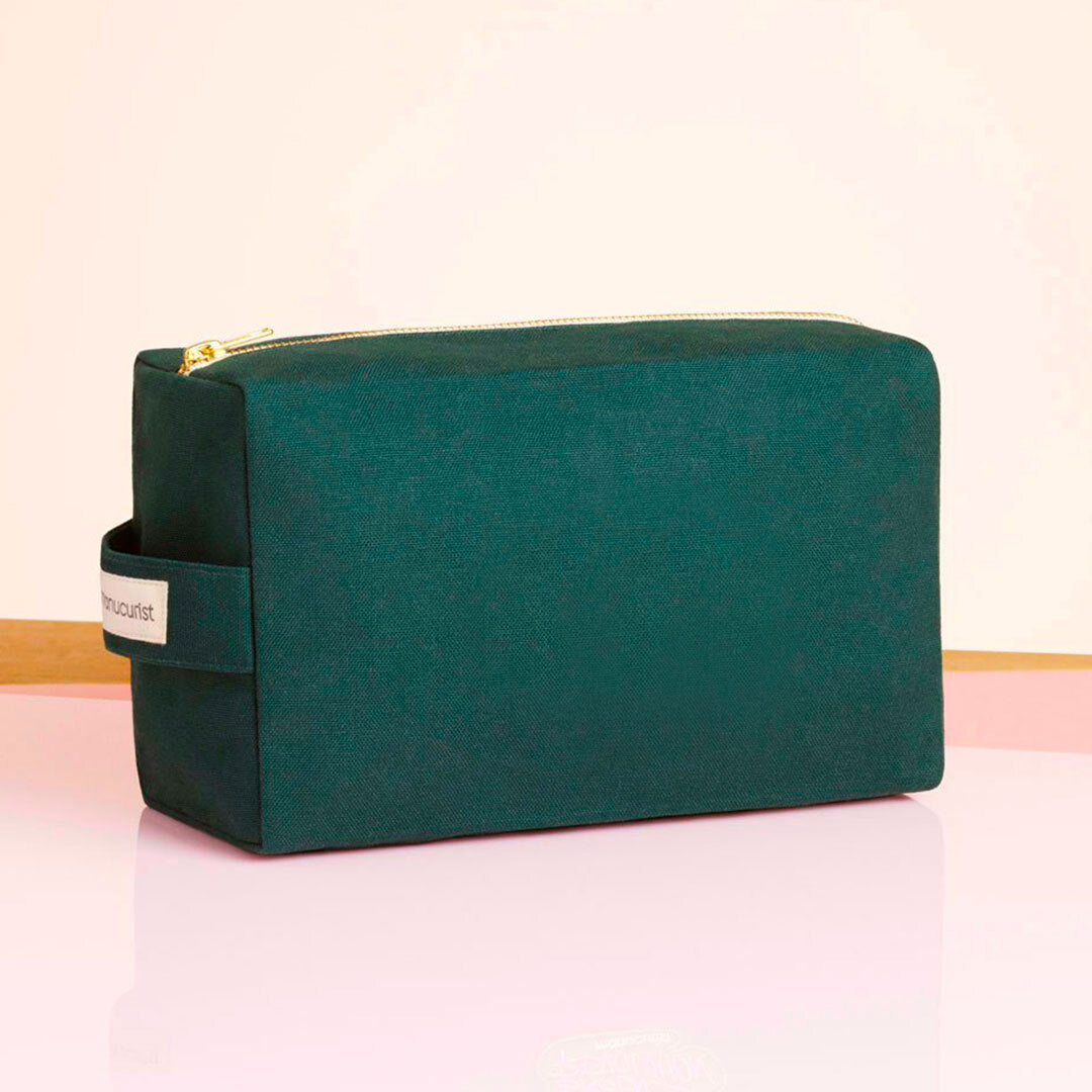 The Large Green Bag by Manucurist-us is a rectangular organic cotton pouch in dark green, featuring a side handle and gold zipper. It is showcased on a glossy pink surface with a light background.