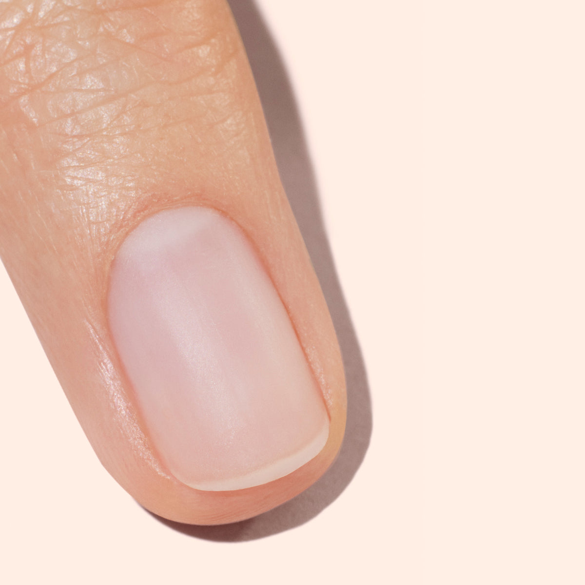 A close-up of a clean, well-groomed fingernail on a finger with smooth, healthy skin, photographed against a light beige background.