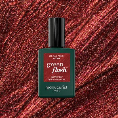 Un flacon de vernis gel Manucurist Utopia se détache sur un fond scintillant rouge profond à paillettes. Le flacon est vert foncé avec un capuchon noir et une étiquette rouge.