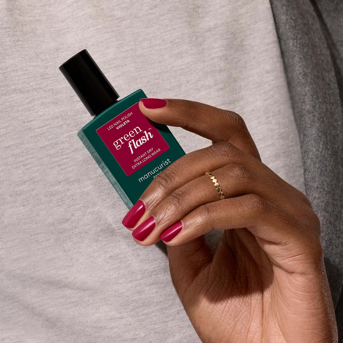 A hand with neatly manicured, deep pink nails holds a bottle of Manucurist Violeta polish against a light gray fabric background.