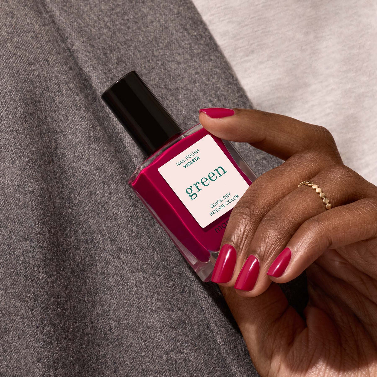 A hand with neatly manicured nails painted in Violeta by Manucurist holds a bottle of the Green™ range nail polish with a black cap against gray and white fabric.