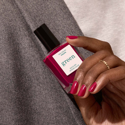 A hand with neatly manicured nails painted in Violeta by Manucurist holds a bottle of the Green™ range nail polish with a black cap against gray and white fabric.