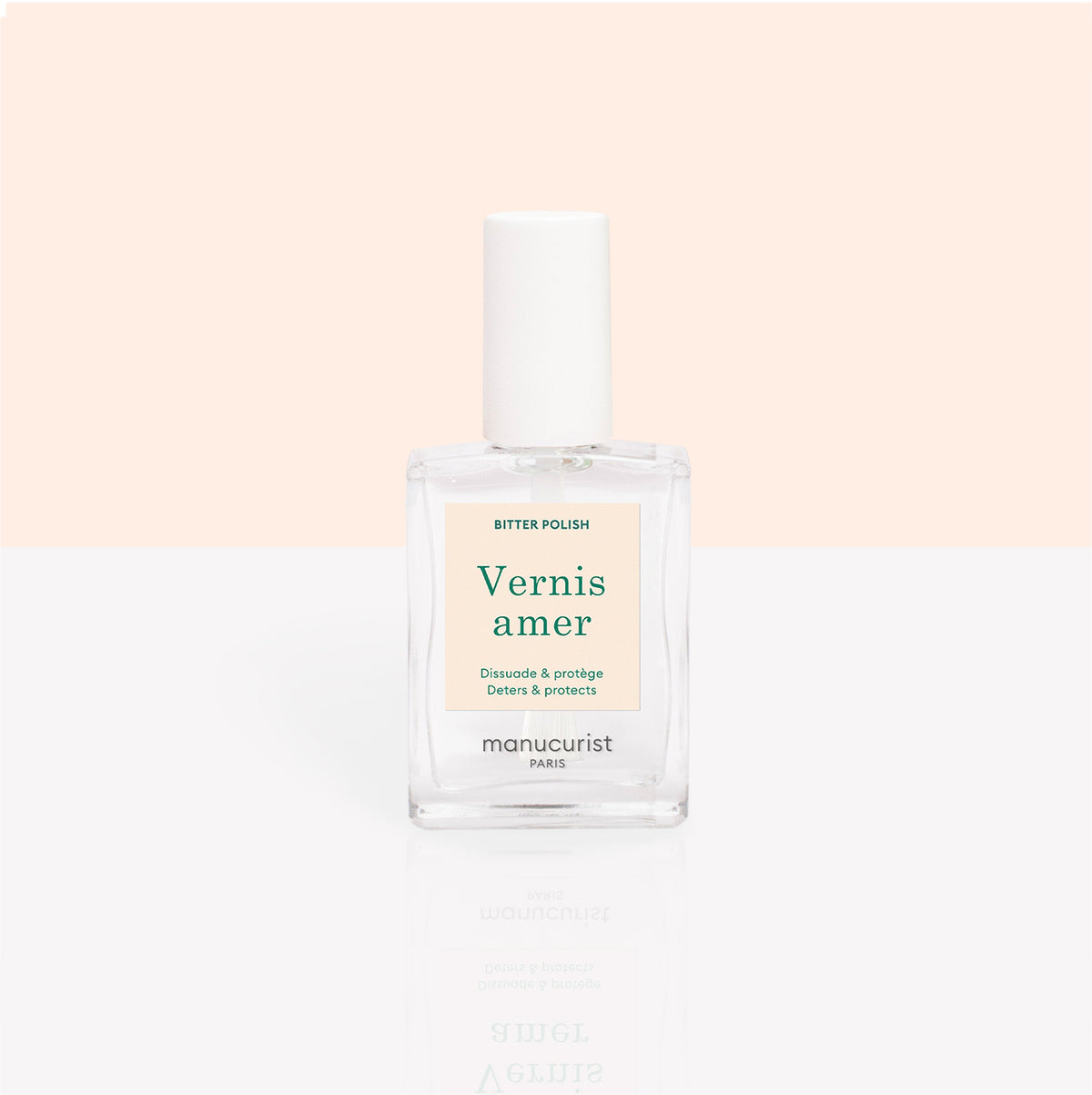A clear bottle of Manucurist Bitter Polish for nail care and to deter nail biting is shown against a soft peach and white background. The label reads "Vernis amer" with green and gold details.