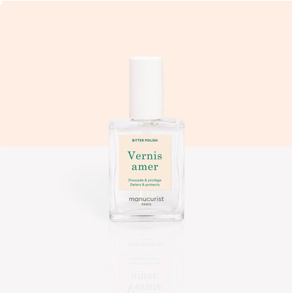 A clear bottle of Manucurist Bitter Polish for nail care and to deter nail biting is shown against a soft peach and white background. The label reads "Vernis amer" with green and gold details.