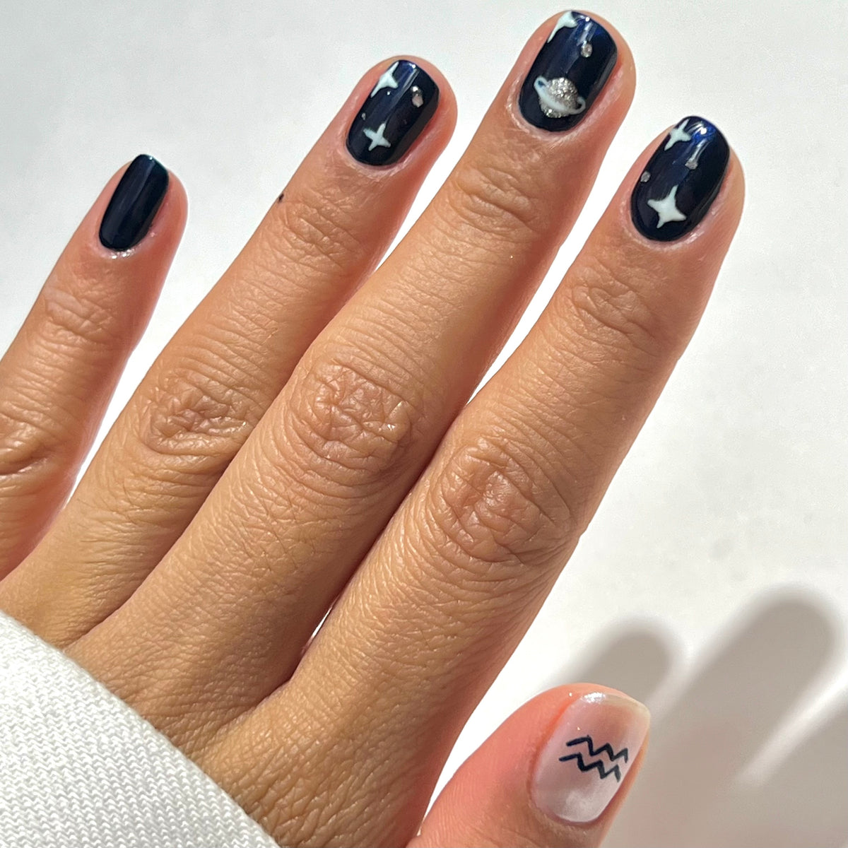 A hand with dark blue nails featuring star and planet designs; the thumb has a light base with a black Aquarius zodiac symbol.