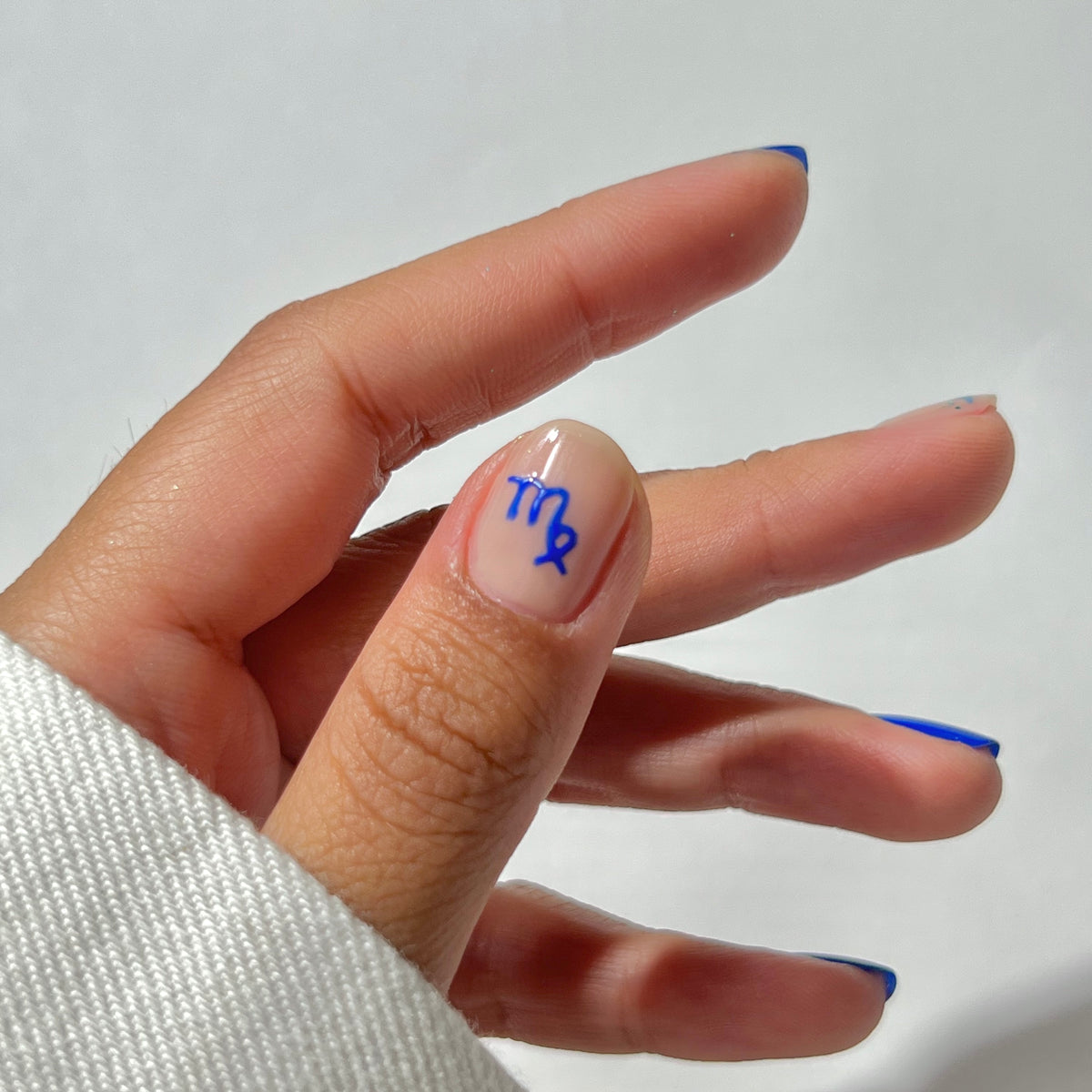 Une main aux ongles soigneusement manucurés, avec un symbole astrologique de la Vierge bleu sur le pouce. Les ongles ont une couleur de base naturelle avec de fines pointes bleues. La main est partiellement recouverte d'une manche blanche sur un fond uni.