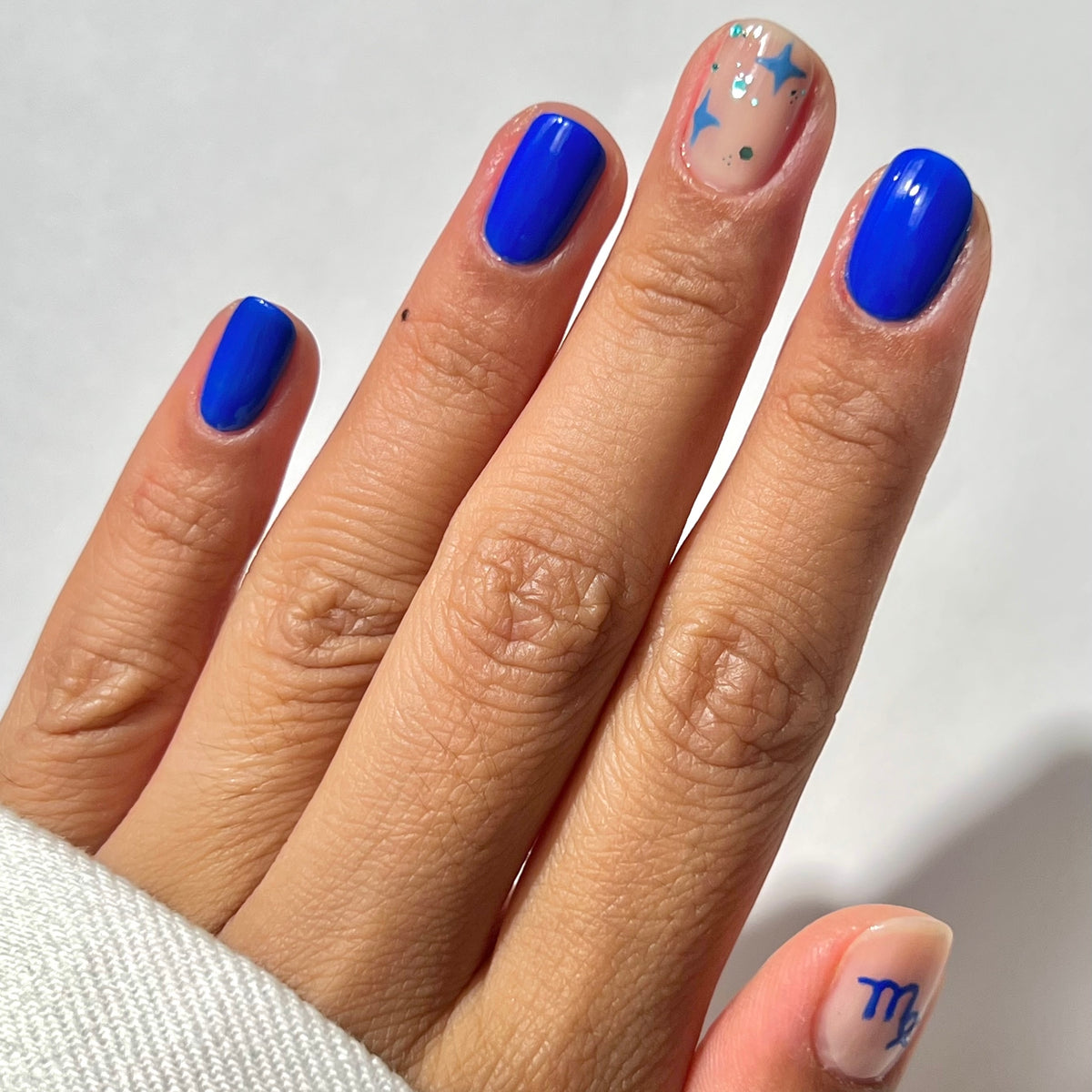 Une main aux ongles vernis de bleu ; l'annulaire présente une base nude avec des formes abstraites bleues et argentées, et l'auriculaire arbore une base nude avec le signe astrologique de la Vierge en bleu.