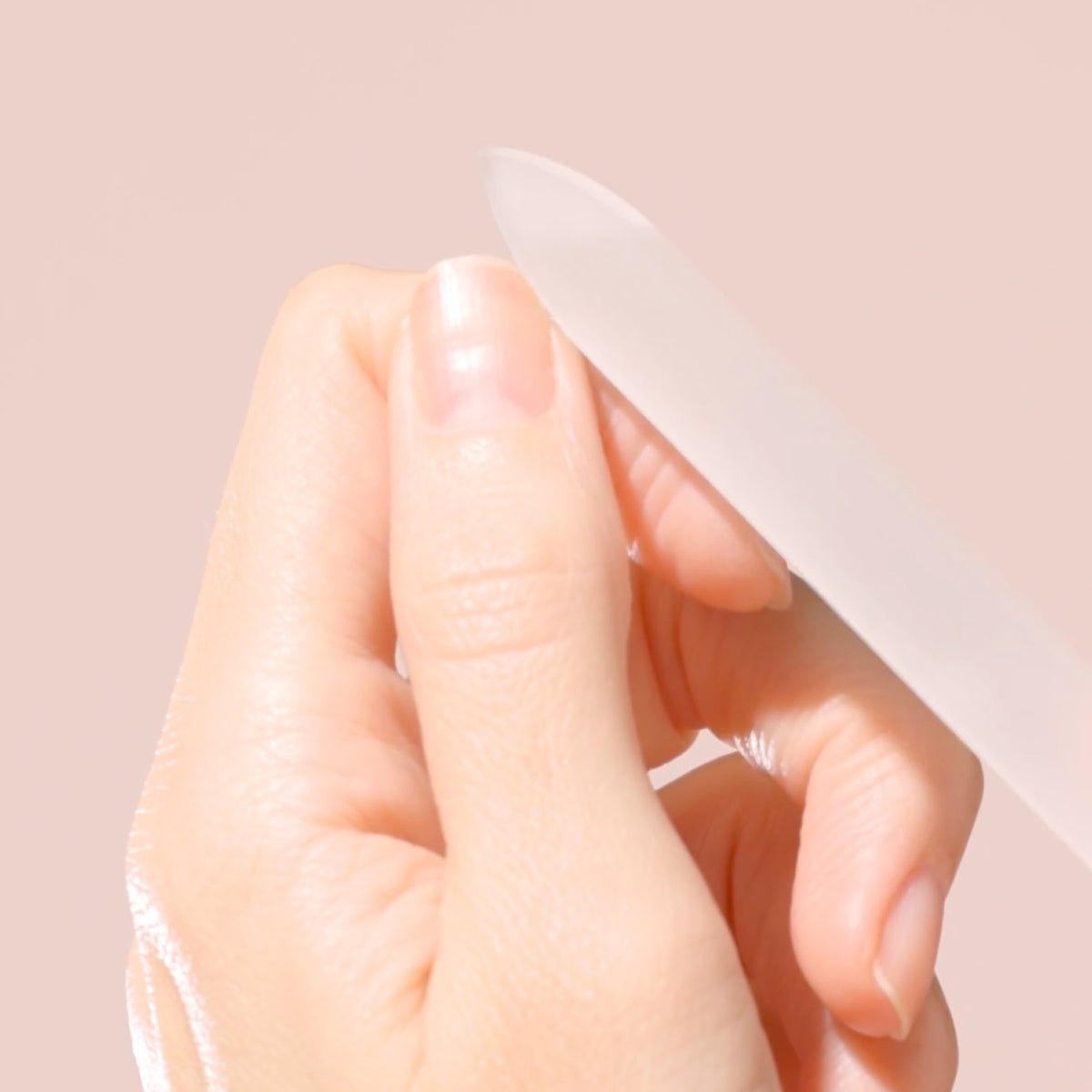 A close-up of a hand using a glass nail file to shape a fingernail, set against a soft pink background.