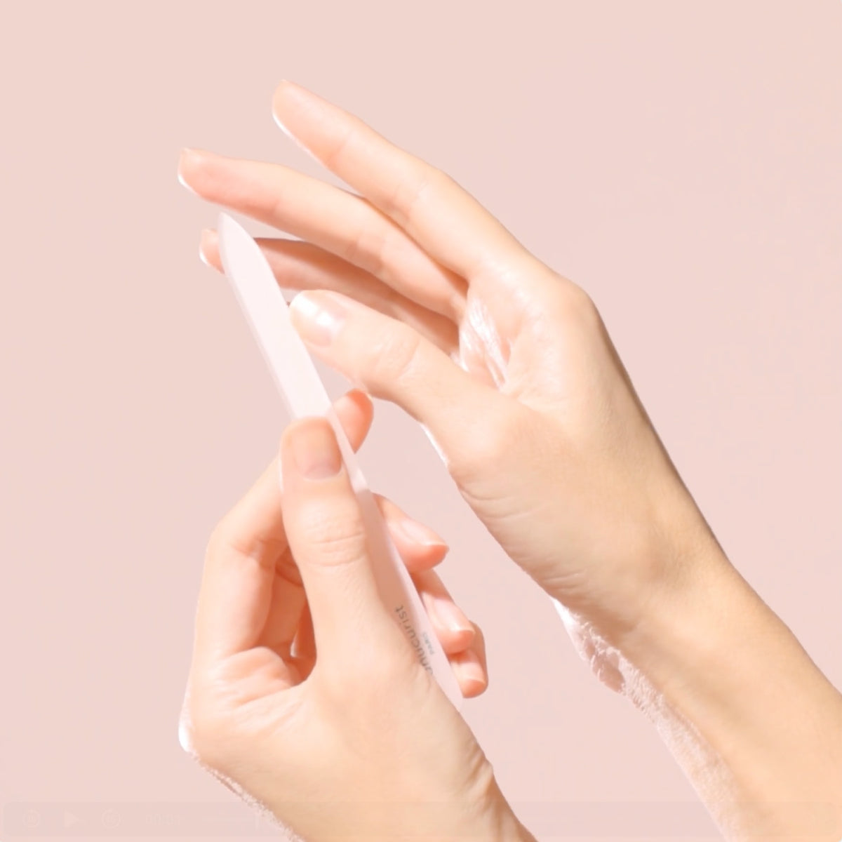 A close-up of two hands, one holding a white nail file and gently filing the nails of the other hand, against a soft pink background.