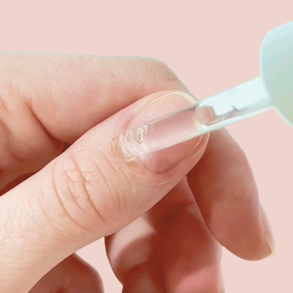 A close-up of a hand applying clear nail serum to a fingernail using a dropper, with a light pink background.