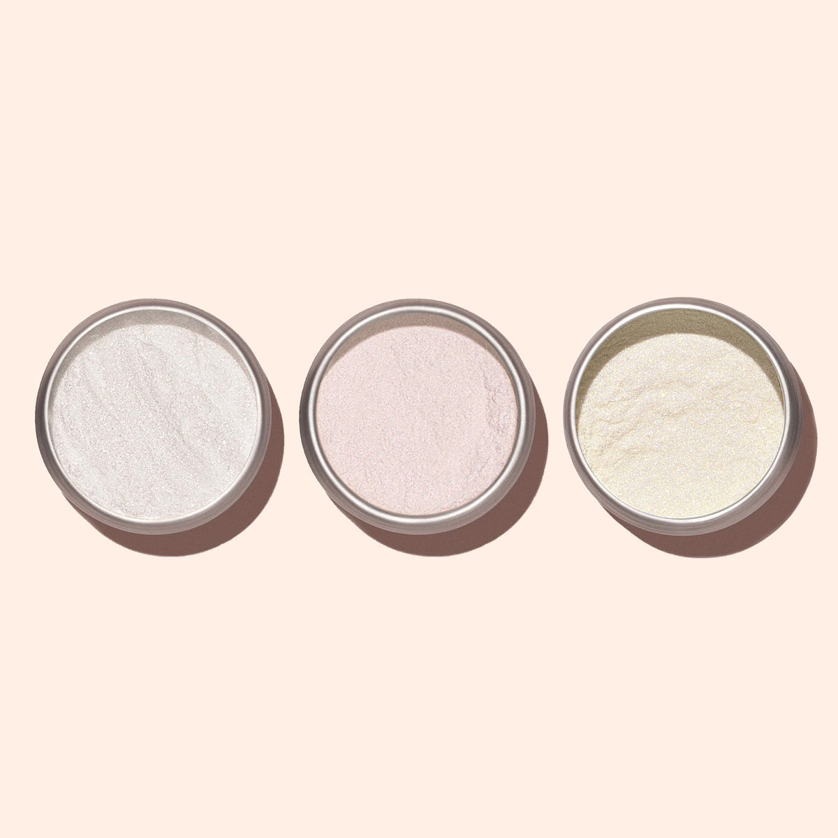 Three small round containers of Manucurist Pink Glazed Effect Powder, in white, light pink, and light yellow shades, are arranged on a pale background, each providing a delicate pearlescent sheen for a subtle glazed finish.
