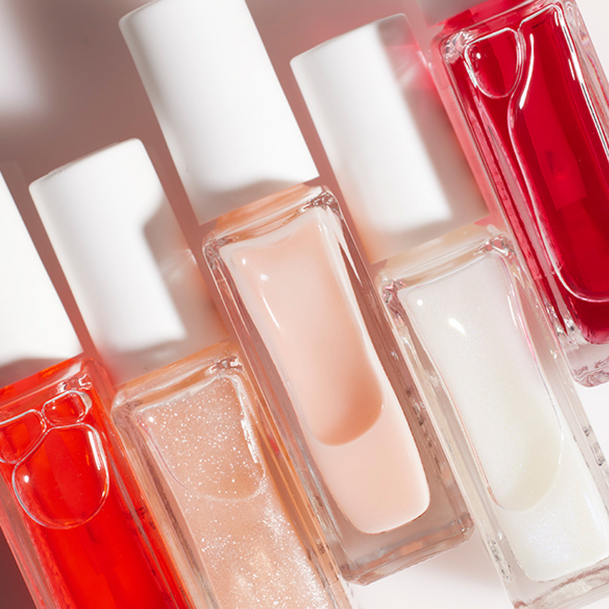 Several glass bottles of nail polish in shades of red, pink, peach, and white with white caps are arranged side by side on a light surface, catching the light and casting soft shadows.