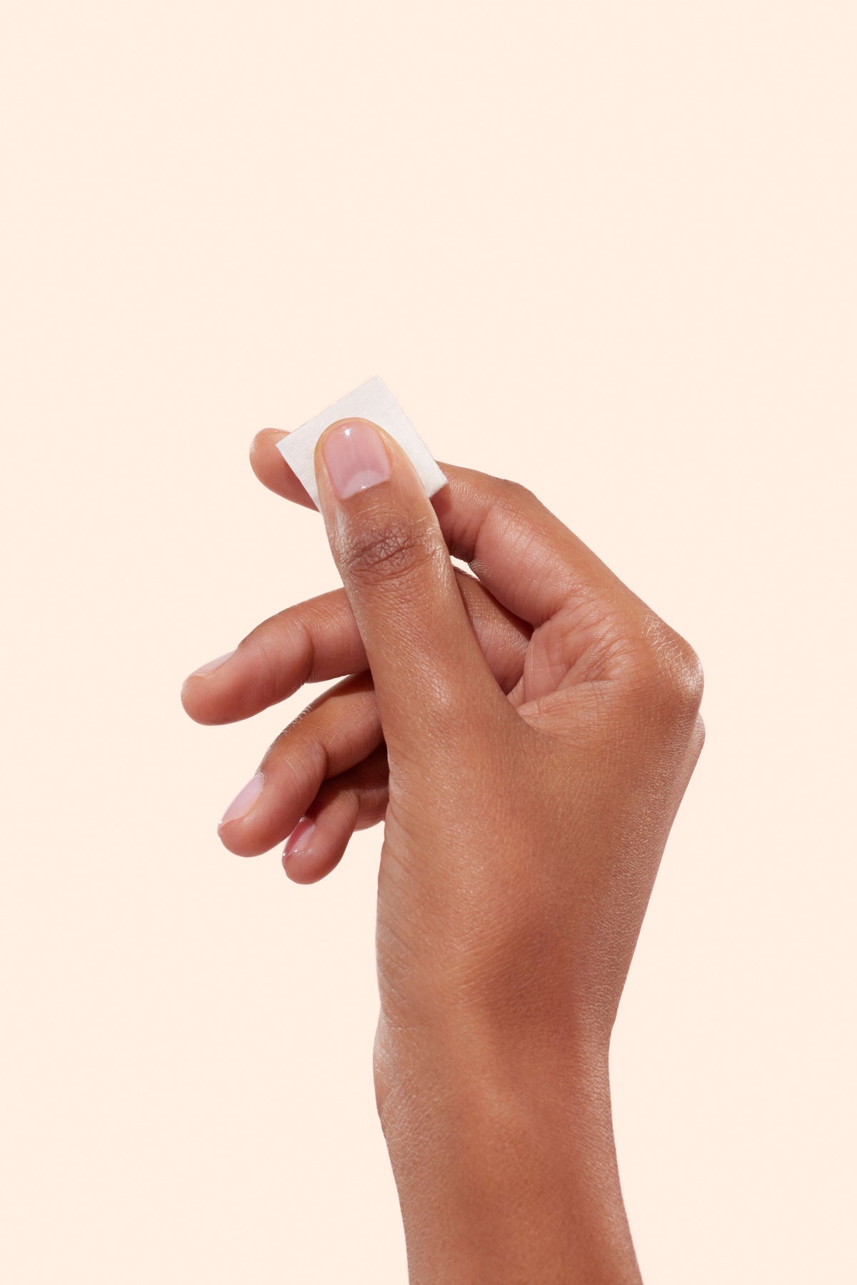 A hand with medium-brown skin holds a small white square, such as a cotton pad or wipe, between the thumb and index finger against a plain light beige background.