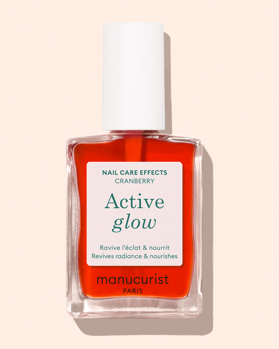 A clear glass bottle of Manucurist Paris “Active Glow” nail care polish with red liquid, a white cap, and a label describing its benefits for reviving radiance and nourishment. The background is light beige.