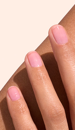 A close-up of a hand with neatly manicured fingernails painted a glossy light pink, resting gently on a persons forearm against a neutral background.