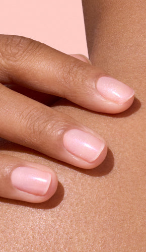 A close-up of a hand with neatly manicured nails painted in a pale, glossy pink shade, resting gently on bare skin with a light brown complexion.