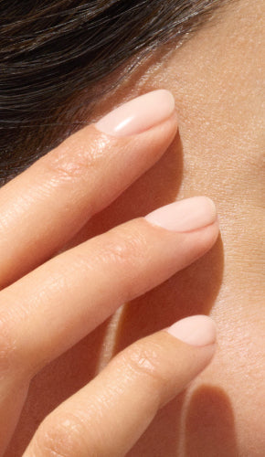 A close-up of a persons fingers with neatly manicured, pale pink nails gently touching their cheek near the temple. The skin appears smooth and well-lit.