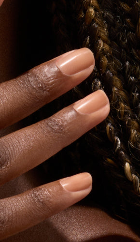A close-up of a hand with medium brown skin tone, showing neatly manicured fingernails painted in a nude shade, positioned near dark brown and blonde braided hair.