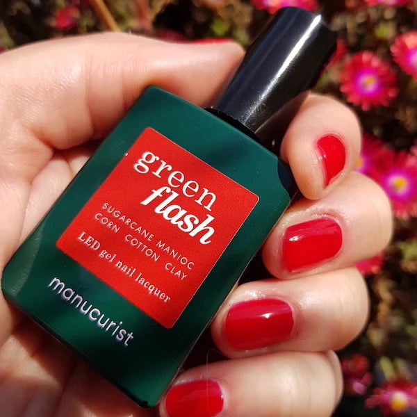 Une main aux ongles peints en rouge vif tient un flacon vert foncé de vernis à ongles gel LED Green Flash™ de Manucurist, sur fond flou de fleurs roses.