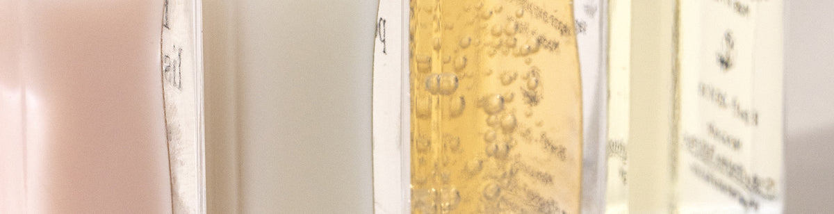 Close-up of three bottles containing liquids in different colors: pink, clear, and yellow. The yellow liquid has visible bubbles. The labels on the bottles are partially visible but not readable.