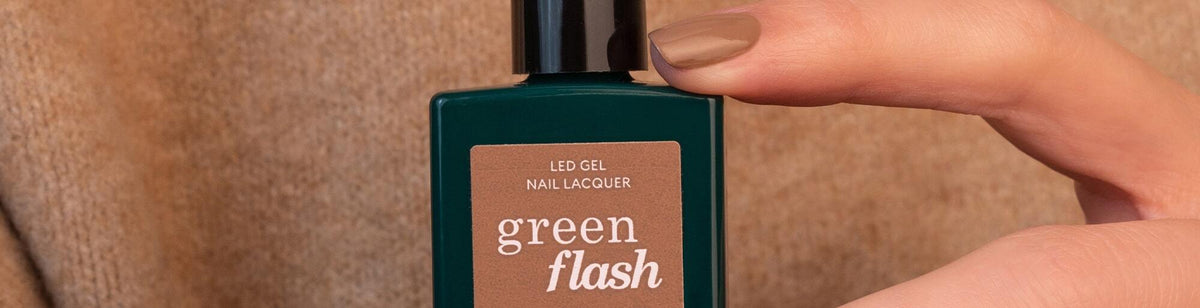 A hand with manicured nails holds a green bottle of green flash LED gel nail lacquer against a beige background.