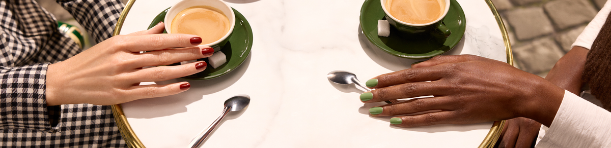 Deux personnes sont assises à une table ronde blanche avec des tasses de café vertes et des cuillères. L’une porte un vernis à ongles rouge et a la peau claire ; l’autre porte un vernis à ongles vert et a la peau foncée. Des morceaux de sucre sont posés sur les soucoupes.