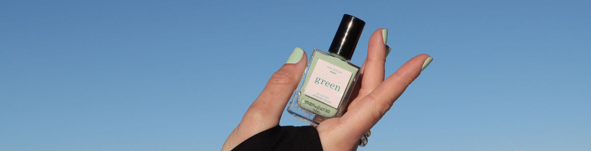 A hand with light green nail polish holds a bottle of green nail polish against a clear blue sky.