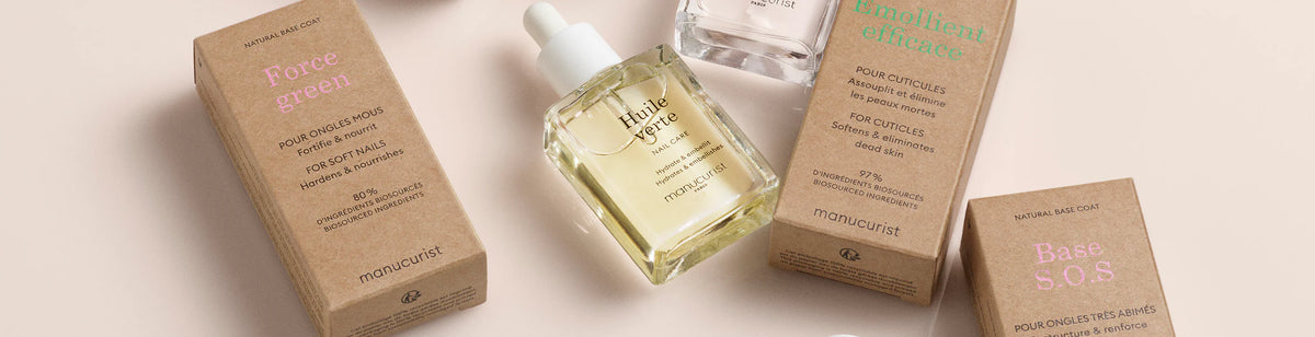 An assortment of Manucurist nail care products, including brown cardboard boxes and glass bottles with droppers, arranged on a light beige surface. Labels show names like “Huile Verte,” “Base S.O.S,” and “Emollient efficace.”.