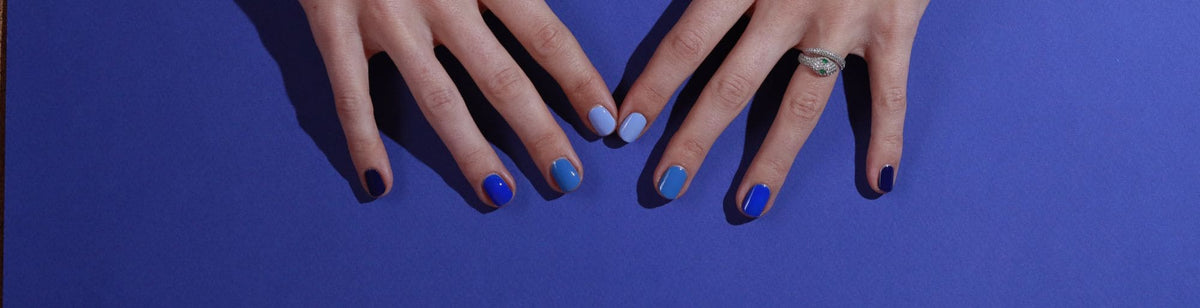 Two hands with manicured nails rest on a blue surface; each fingernail is painted a different shade of blue, and one hand has a ring on the ring finger.