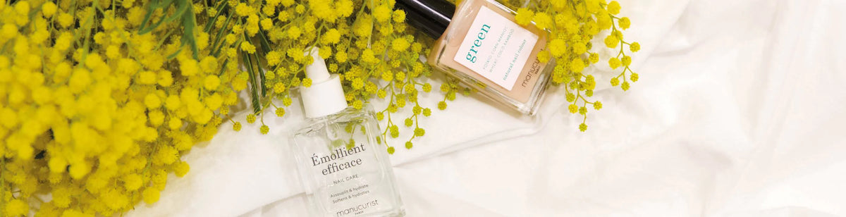 Two glass perfume bottles lie on a white fabric surrounded by sprigs of small, bright yellow flowers. One bottle is labeled “green” and the other reads “Emollient efficace.”.