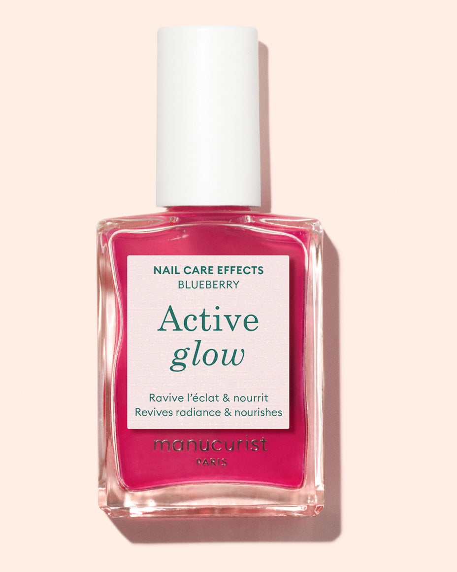 A bottle of Manucurist Paris Active Glow nail polish with a white cap, containing bright pink liquid and labeled as Nail Care Effects Blueberry for reviving radiance and nourishing nails.
