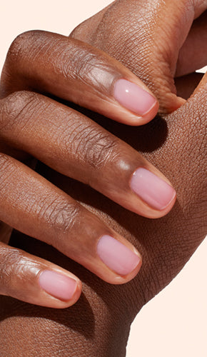 A close-up of a hand with neatly manicured nails painted in a soft, pale pink shade, resting gently on the wrist of the other hand against a light background.