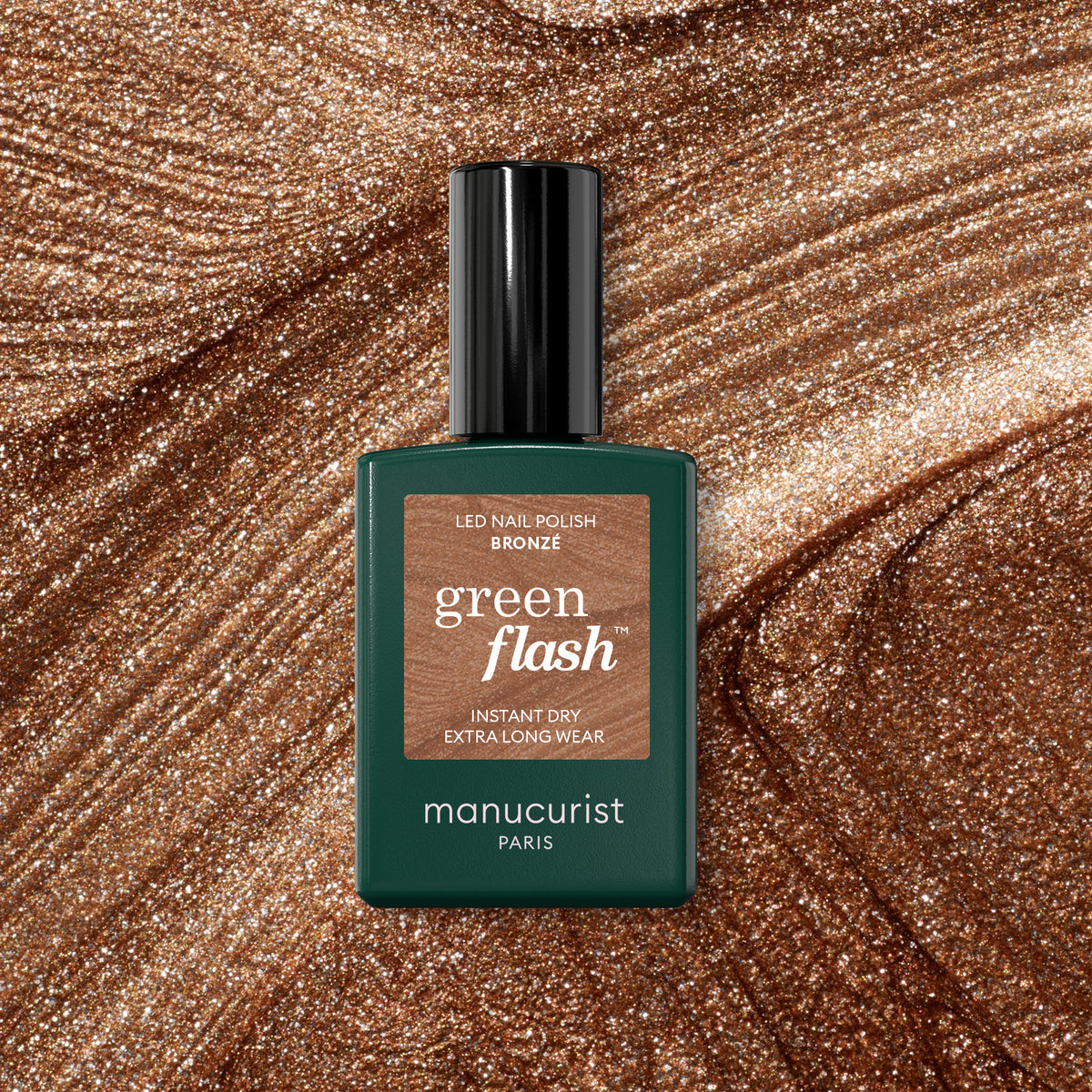 A bottle of Manucurist Bronzé Green Flash gel polish rests on a shimmery bronze background, perfectly matching the dazzling shade of this nail polish.