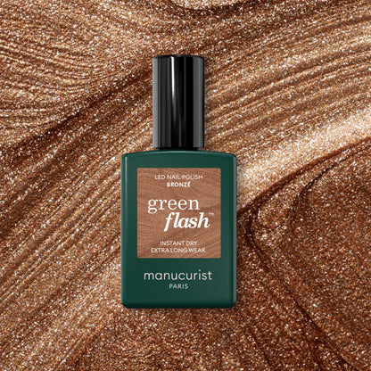 A bottle of Manucurist Bronzé Green Flash gel polish rests on a shimmery bronze background, perfectly matching the dazzling shade of this nail polish.